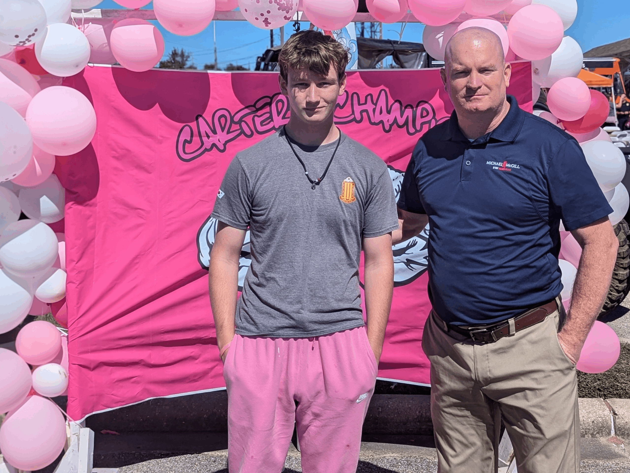 Michael McGill and his son are standing in front of a pink backdrop decorated with white and pink balloons. The backdrop has the text "CARTERS CHAMPIONS". Michael McGill III on the left is wearing a gray t-shirt and pink pants, while the Michael McGill on the right is wearing a navy blue polo shirt with a the word Michael McGill and Stay American underneath the name and khaki pants. The setting appears to be at the Pink Dress Run in Gulfport, MS