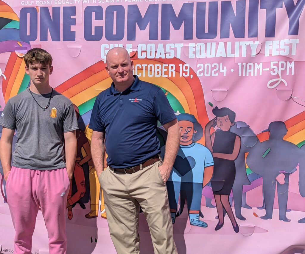 Michael McGill and his son stand infront of the One Community sign at the Gulf Coast Equality Fest 2024 in Biloxi, MS