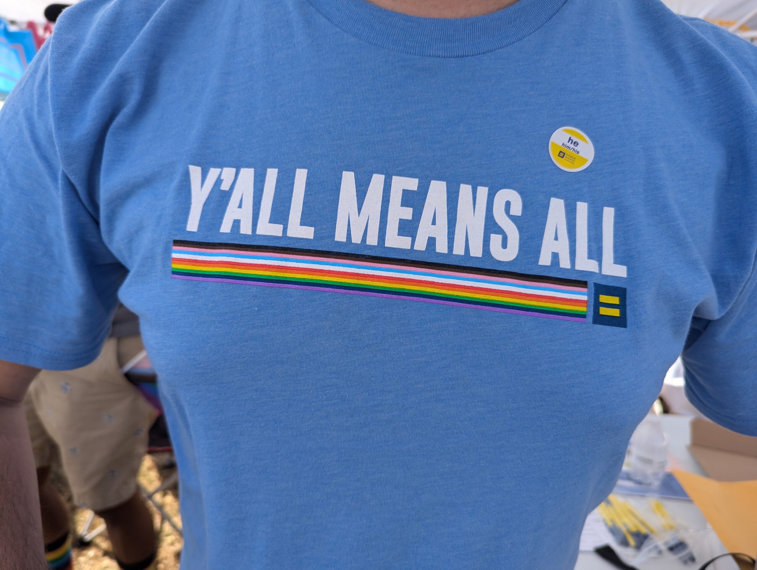 Y'all Means All written in white letters on a blue shirt at the Gulf Coast Equality Fest 2024.