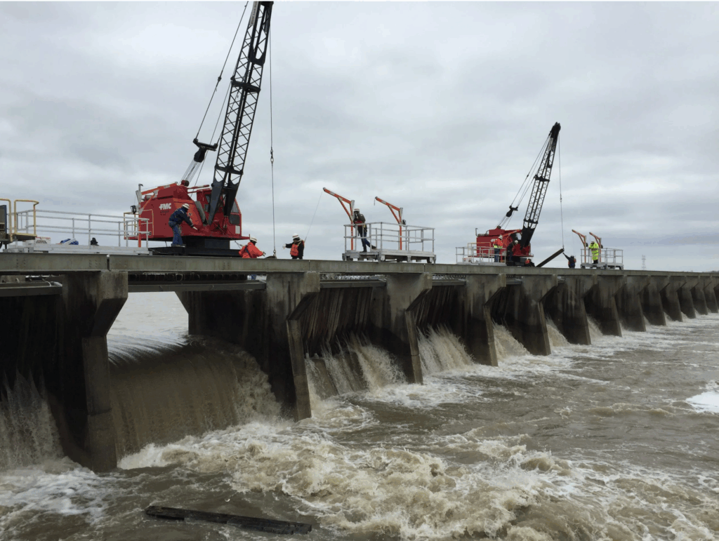 Workers perform maintenance and monitoring operations atop the Bonnet Carré Spillway in Louisiana, with water flowing below and a cargo ship passing in the background.