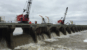 Workers perform maintenance and monitoring operations atop the Bonnet Carré Spillway in Louisiana, with water flowing below and a cargo ship passing in the background.