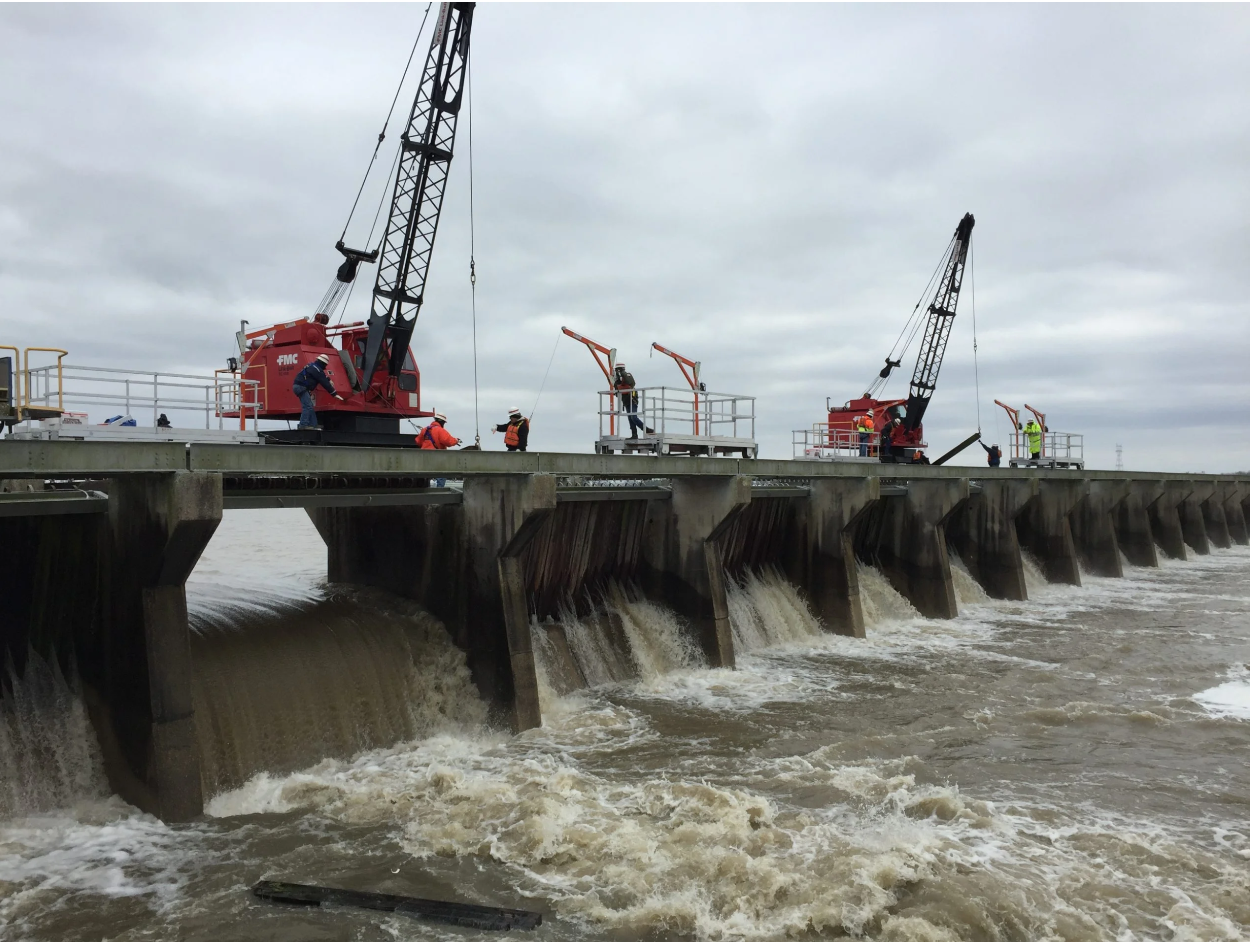 Workers perform maintenance and monitoring operations atop the Bonnet Carré Spillway in Louisiana, with water flowing below and a cargo ship passing in the background.