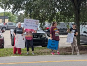 Protesters Four individuals stand peacefully under a tree outside the Gulfport Chamber of Commerce’s Legislative Luncheon, holding handmade signs that read messages like “Defend the Constitution” and “We the People.” They are members of the Mississippi Gulf Coast Mutual Aid Collective, publicly expressing their concerns about government, constitutional rights, and fiscal responsibility.