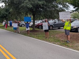 PXL_20250805_160912467 Members of the Mississippi Gulf Coast Mutual Aid Collective line the roadside outside the Gulfport Chamber of Commerce’s Legislative Luncheon, holding handmade protest signs with messages about fiscal responsibility, veterans’ support for the Constitution, and the defense of democracy. The peaceful demonstrators stand spaced under the trees, expressing their civic concerns to passing attendees.