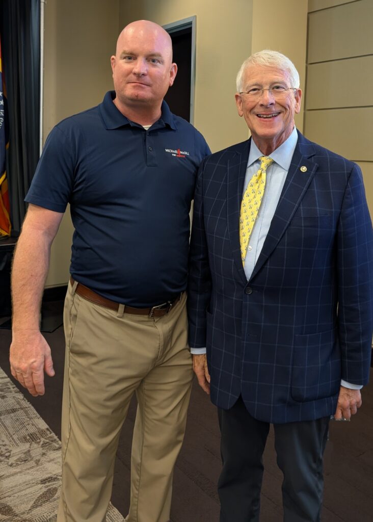 PXL_20250805_180046281~3 Michael McGill stands beside U.S. Senator Roger Wicker at the 2025 Gulfport Chamber Legislative Luncheon. McGill wears a navy polo shirt with his campaign logo, while Senator Wicker is dressed in a checkered navy blazer and yellow tie, both smiling for the photo.