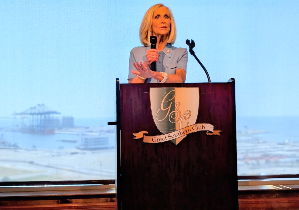 Mississippi Attorney General Lynn Fitch speaks at a podium during the Harrison County GOP Luncheon at the Great Southern Club, with the Gulfport port visible in the background.
