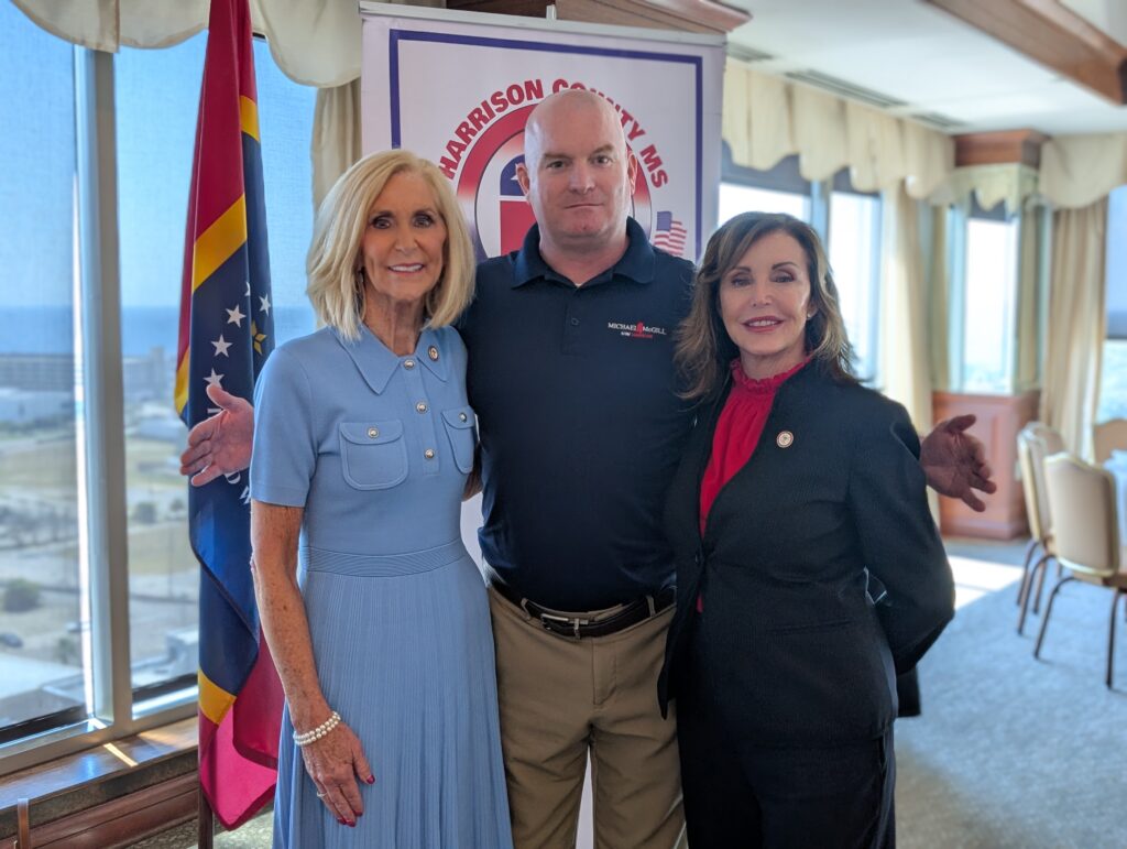 Michael McGill stands with Mississippi Attorney General Lynn Fitch and Starr Chapman at the Harrison County GOP Luncheon, posing in front of a banner and state flag.