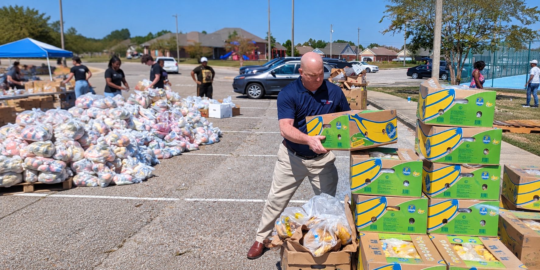 Michael McGill carrying a box of bananas at a community food distribution while volunteers sort bags of food in a parking lot