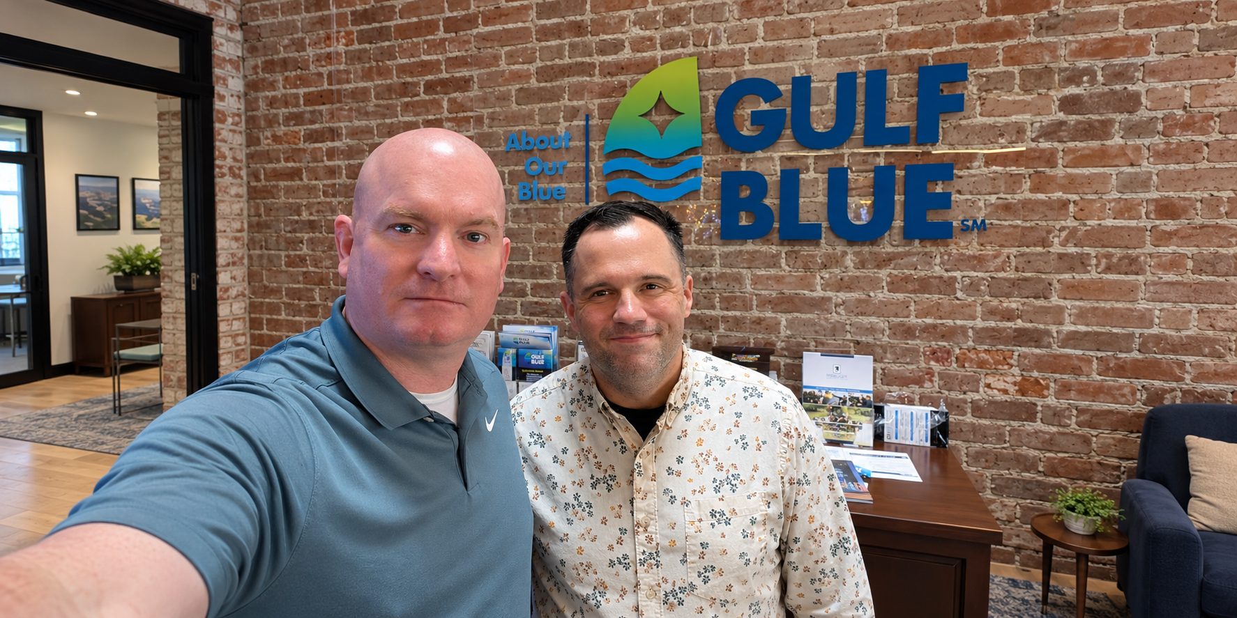 Michael McGill and another man smiling for a selfie in front of the Gulf Blue sign on a brick wall